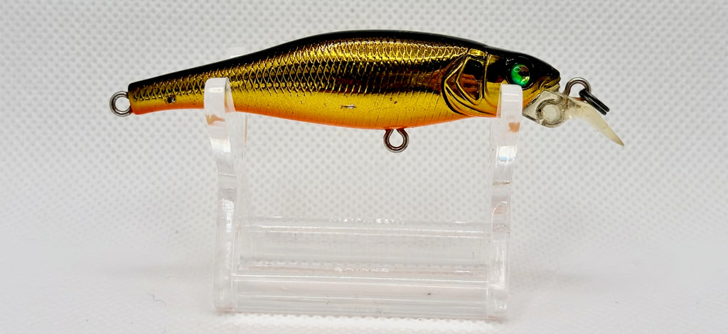 A gold and black metallic fishing lure with a clear plastic diving lip, displayed on a clear acrylic stand against a white textured background.