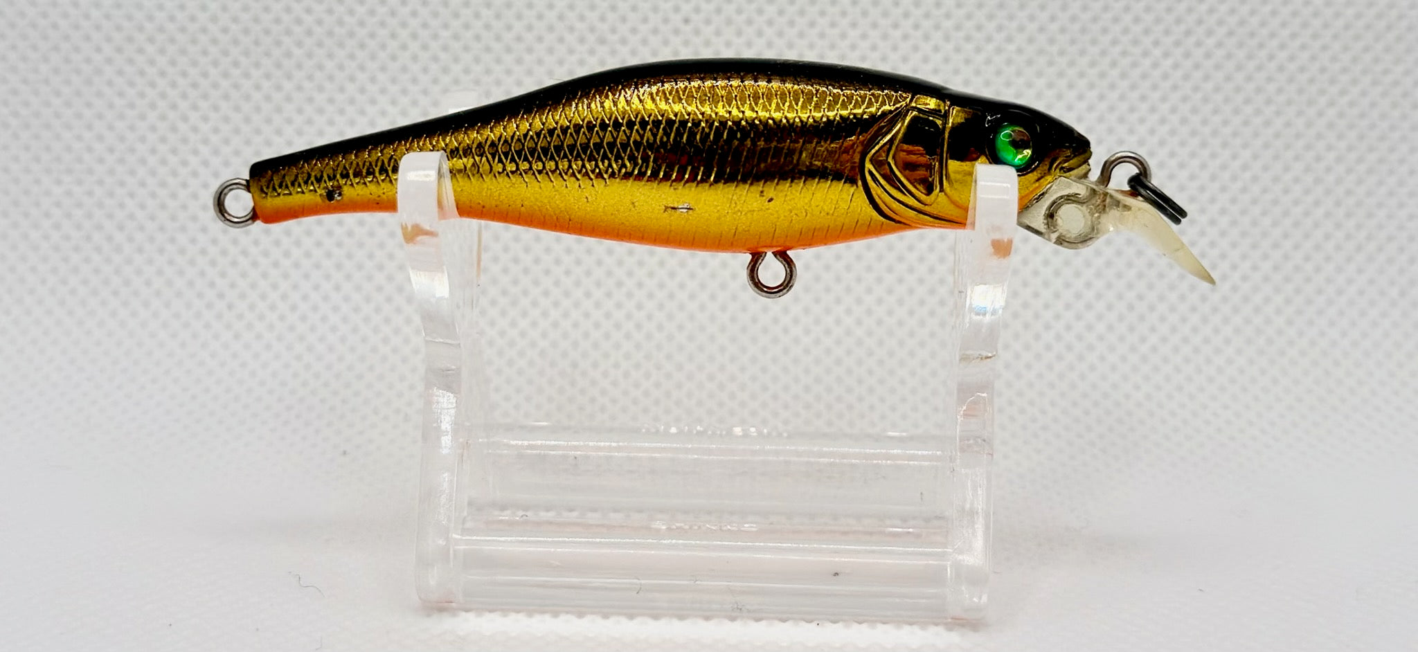 A gold and black metallic fishing lure with a clear plastic diving lip, displayed on a clear acrylic stand against a white textured background.