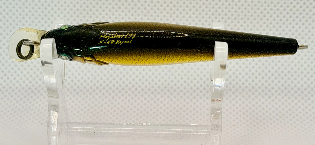 A fishing lure displayed on clear acrylic stands against a white textured background. The lure has a metallic gold and black body with a green-blue iridescent edge and a white bail.
