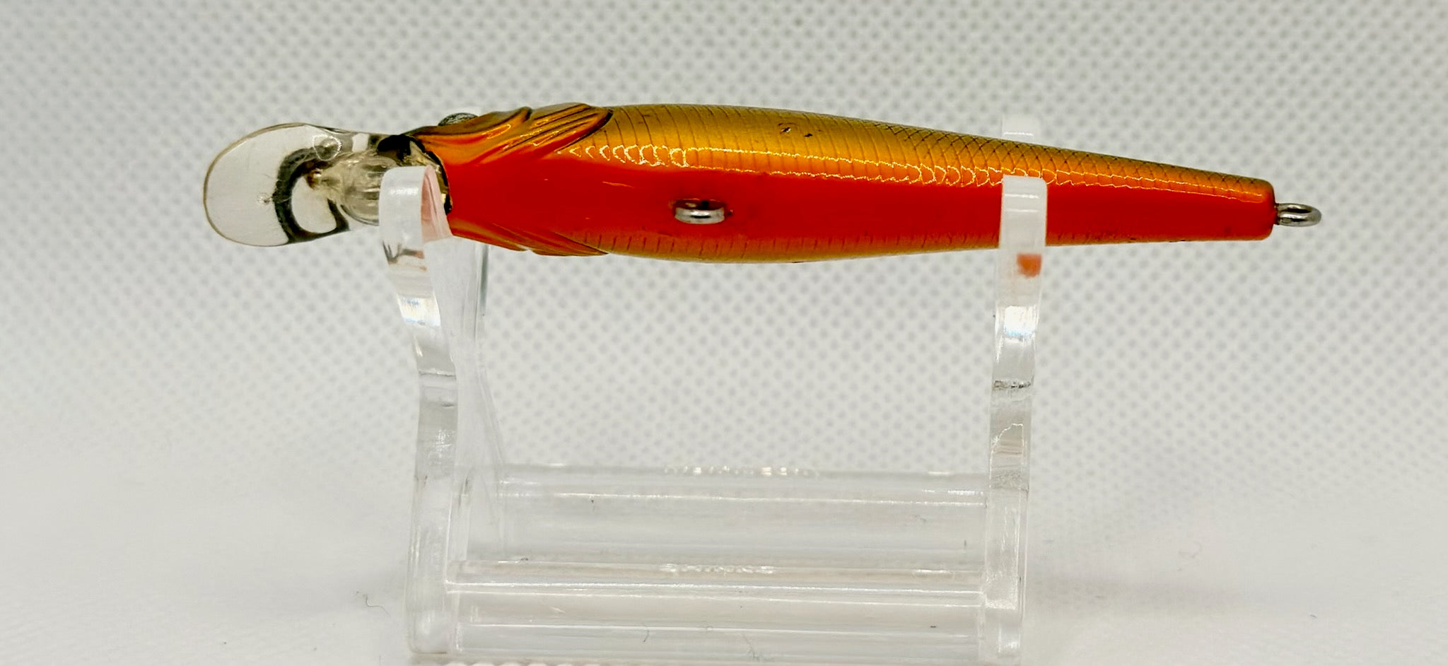 A fishing lure with a metallic gold and red body, mounted on a clear plastic display stand against a white textured background.