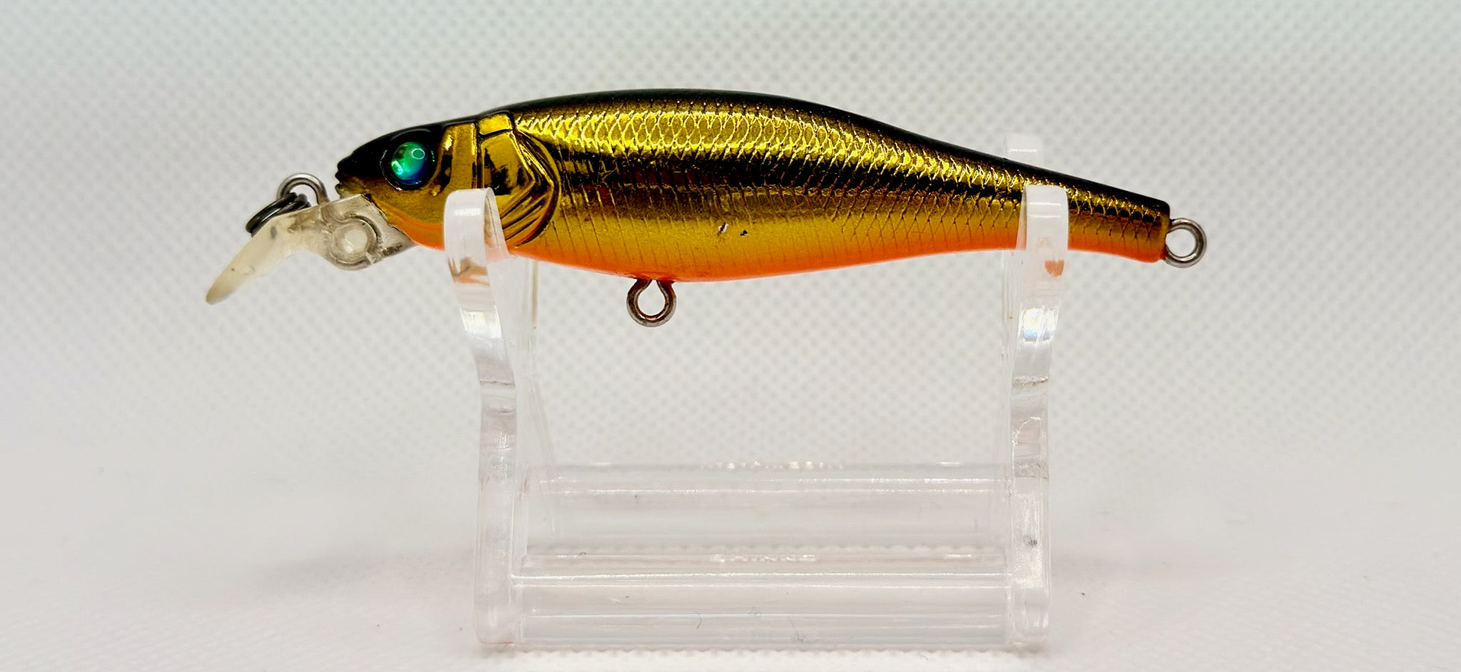 A metallic gold and orange fishing lure with a clear diving lip, displayed on a clear acrylic stand against a white textured background.
